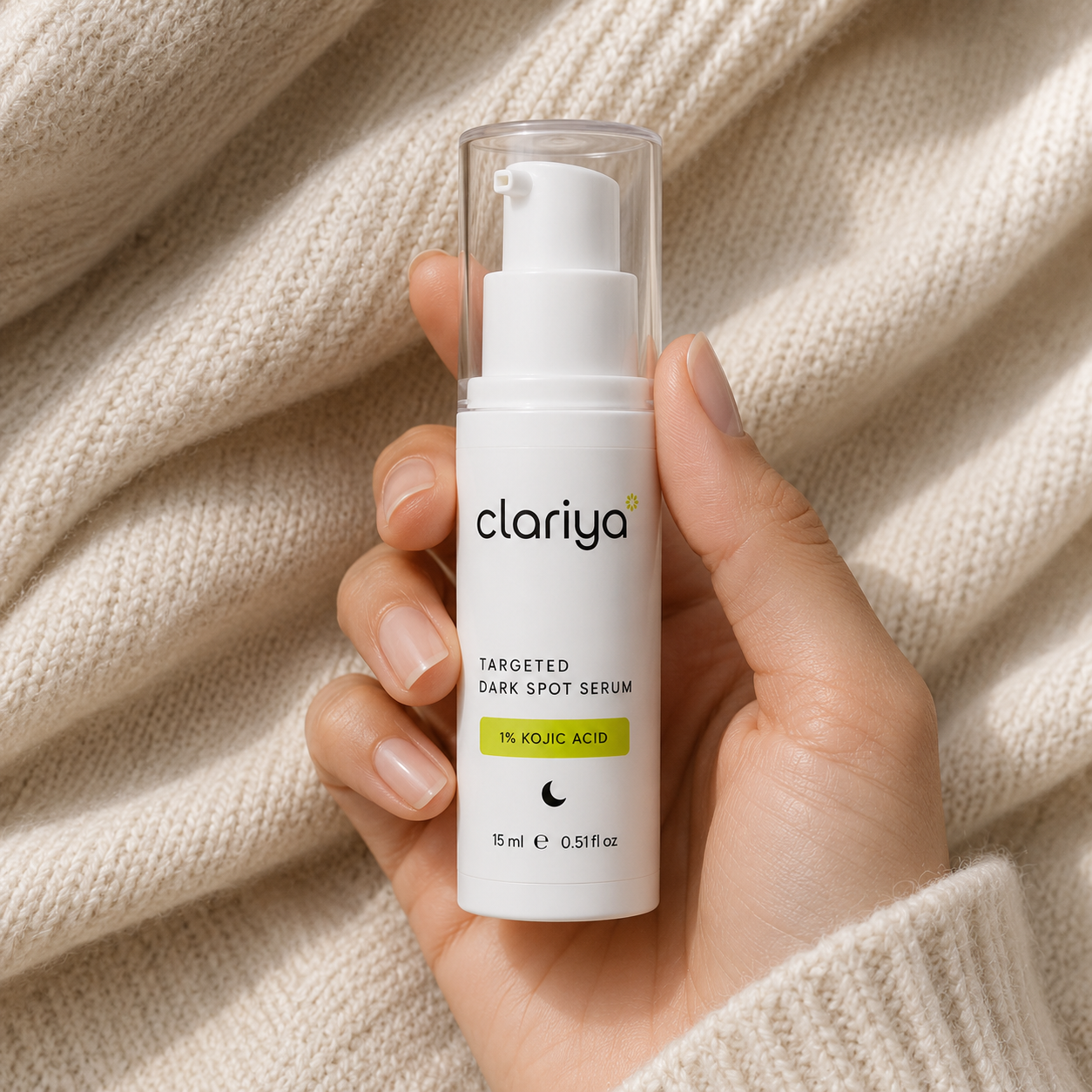 A hand with even-toned skin holding the white CLARIYA Targeted Dark Spot Serum bottle against soft oatmeal knit fabric.
