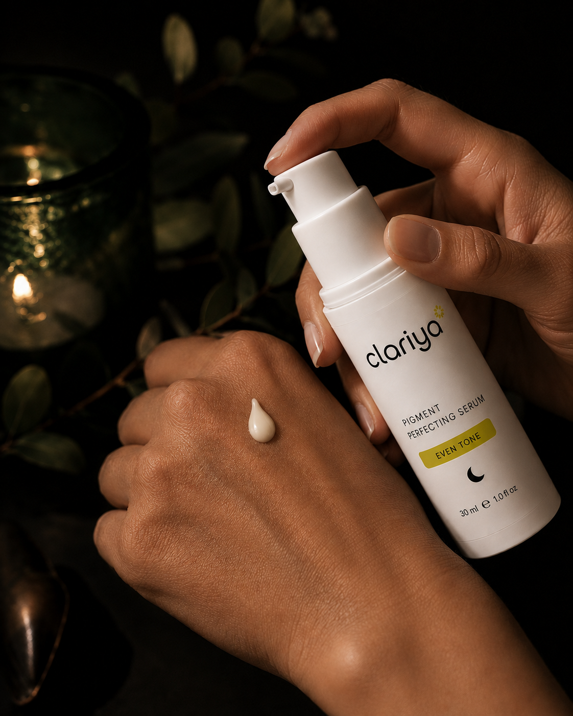 Hands pressing the CLARIYA Pigment Perfecting Serum pump with a serum drop on the back of the hand, a candle in green glass glowing in the dark evening background.