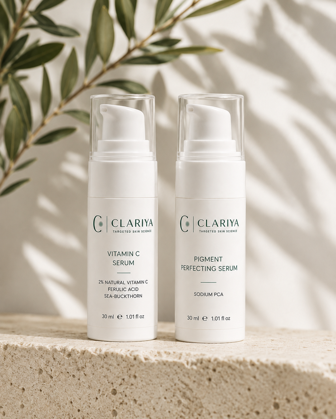 The CLARIYA Vitamin C Serum and Pigment Perfecting Serum standing side by side on a travertine ledge with an olive branch and soft daylight shadows.
